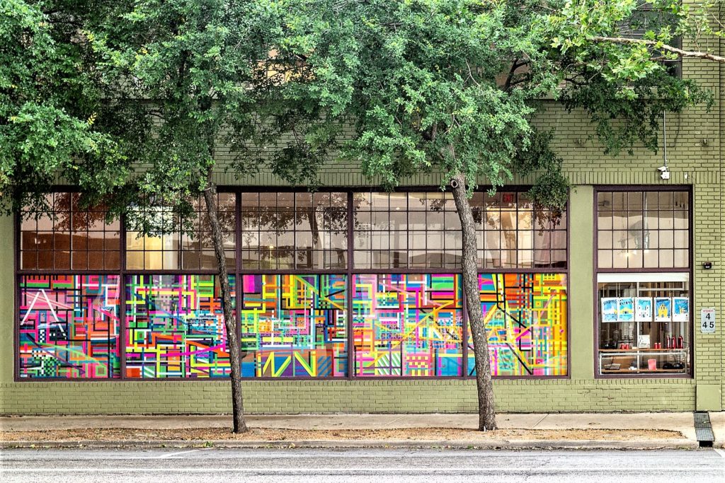 a city-view shot of a gallery building with light green walls, the windows of the building are filled with vivid drawings of colorful lines and shades